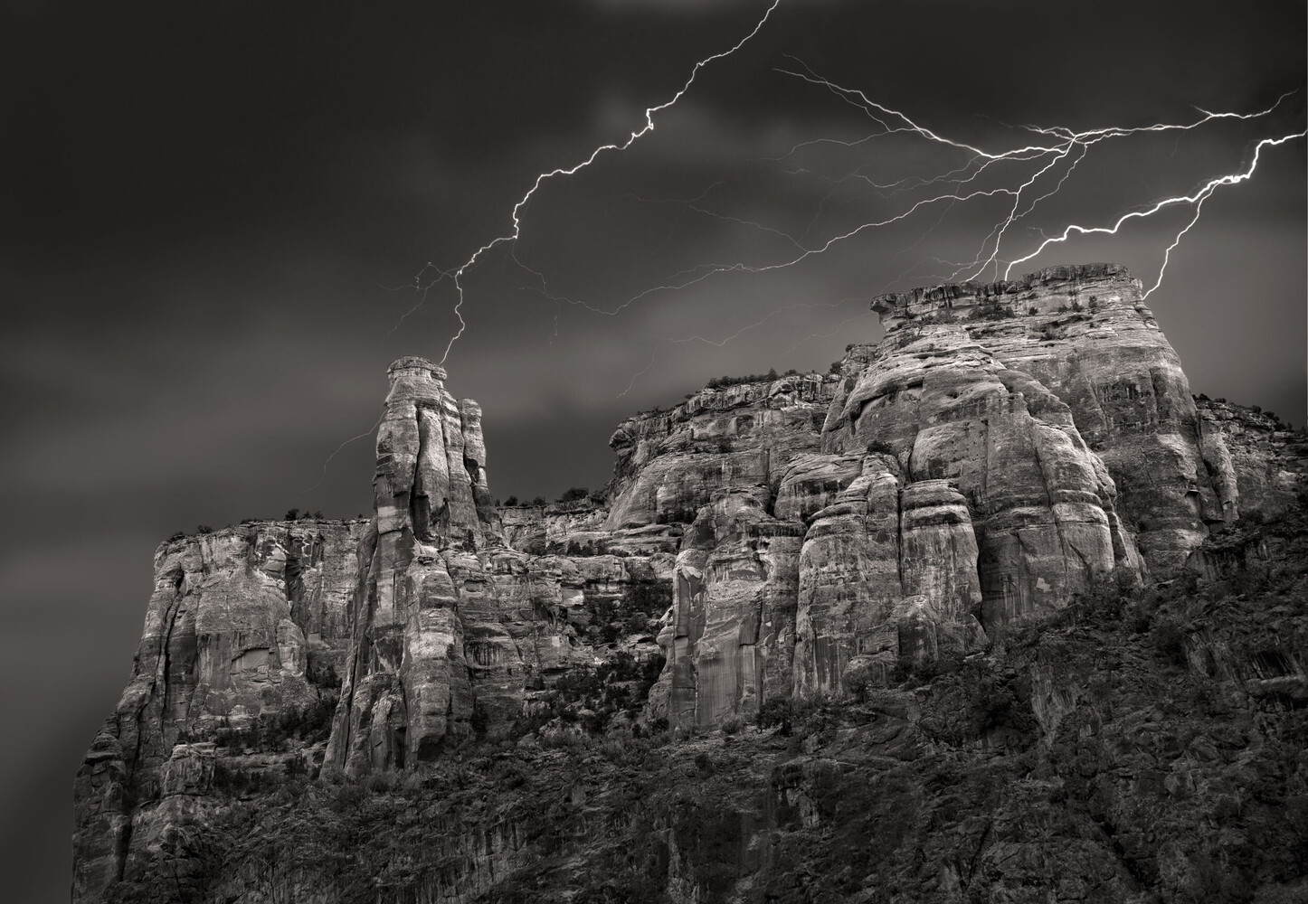Lightning bolts striking behind a sandstone butte and rock spire under a dark, stormy sky, in black and white.