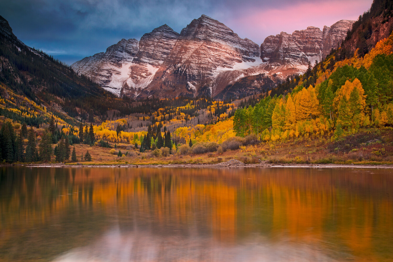 Snow-streaked red mountain peaks rise above a still lake that reflects golden autumn aspen trees and a pink evening sky.
