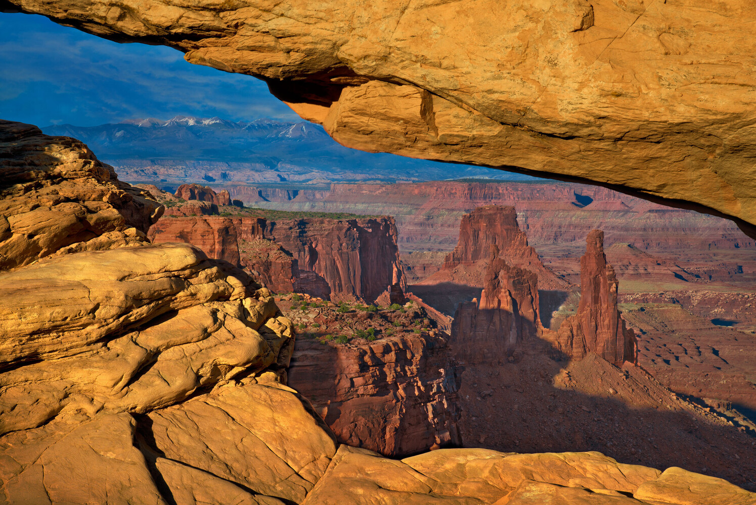 Sandstone natural arch glowing gold above red rock canyons and spires, with snowy mountains on the horizon