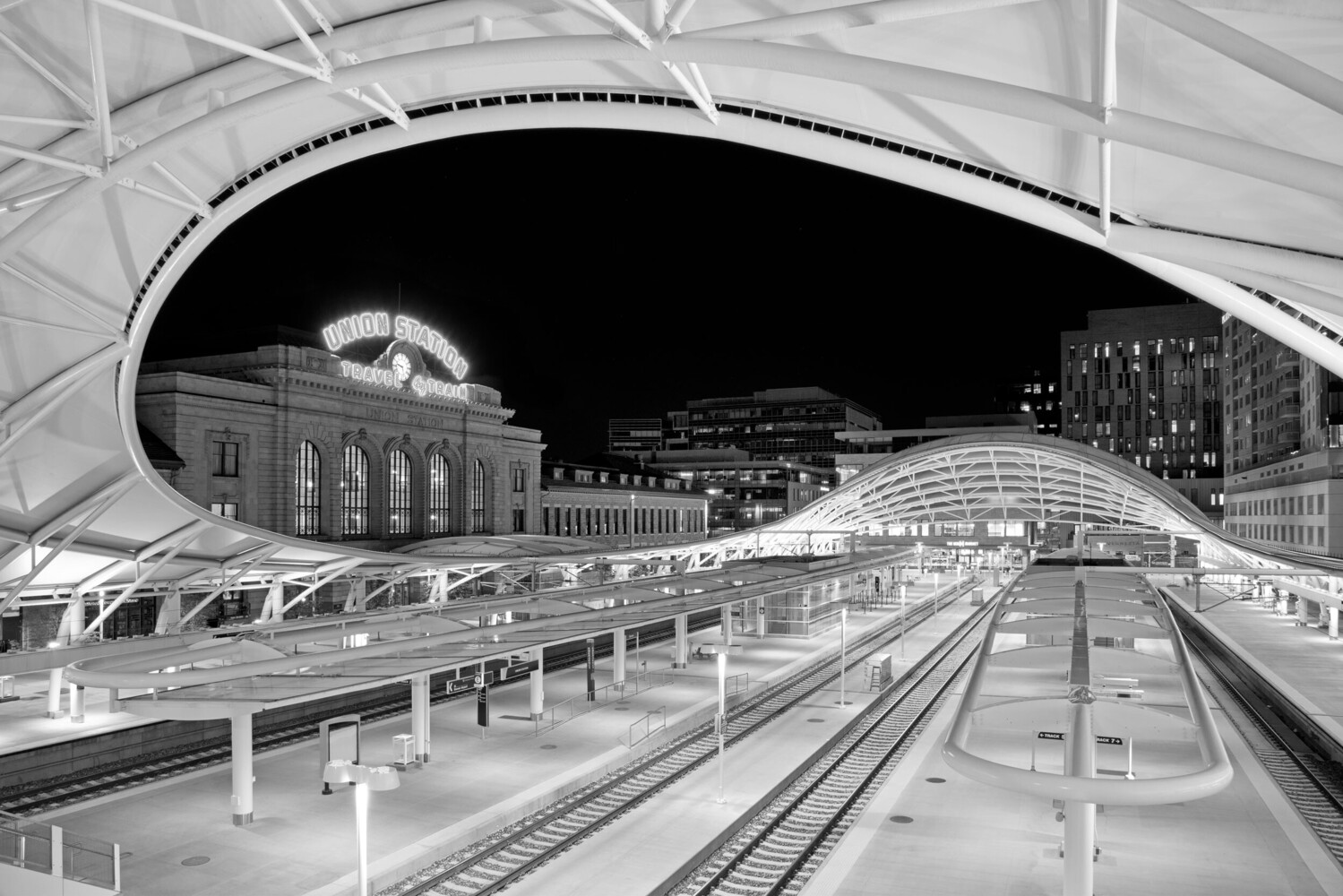 Black and white night view of Union Station's lit sign and curved white canopy over empty train platforms and tracks.