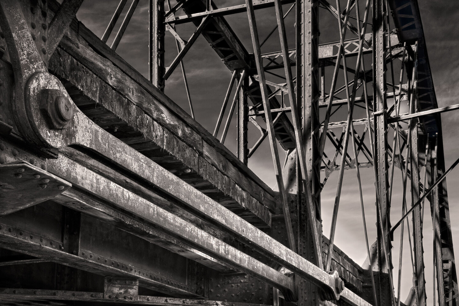 Black and white low-angle view of rusted steel bridge girders and riveted lattice trusses against a cloudy sky