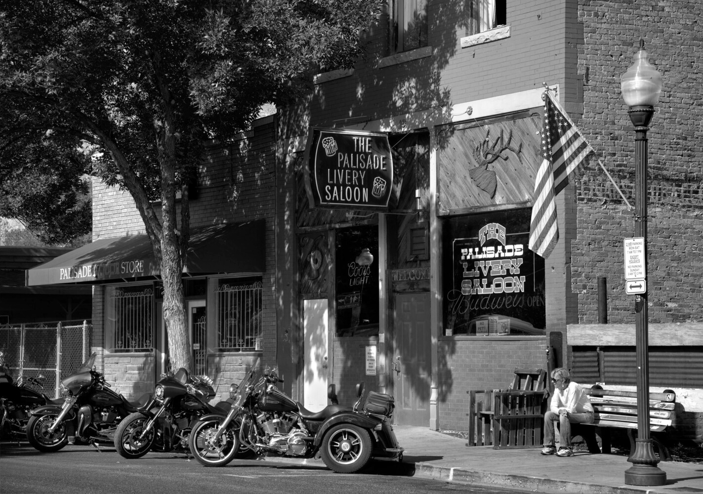 Black and white street scene of motorcycles parked outside The Palisade Livery Saloon, with a seated man on a bench at right.