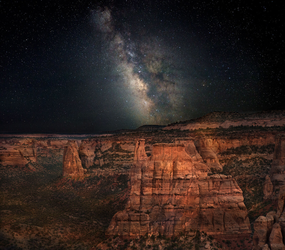 Milky Way galaxy arching over glowing red sandstone spires and canyon walls at night.