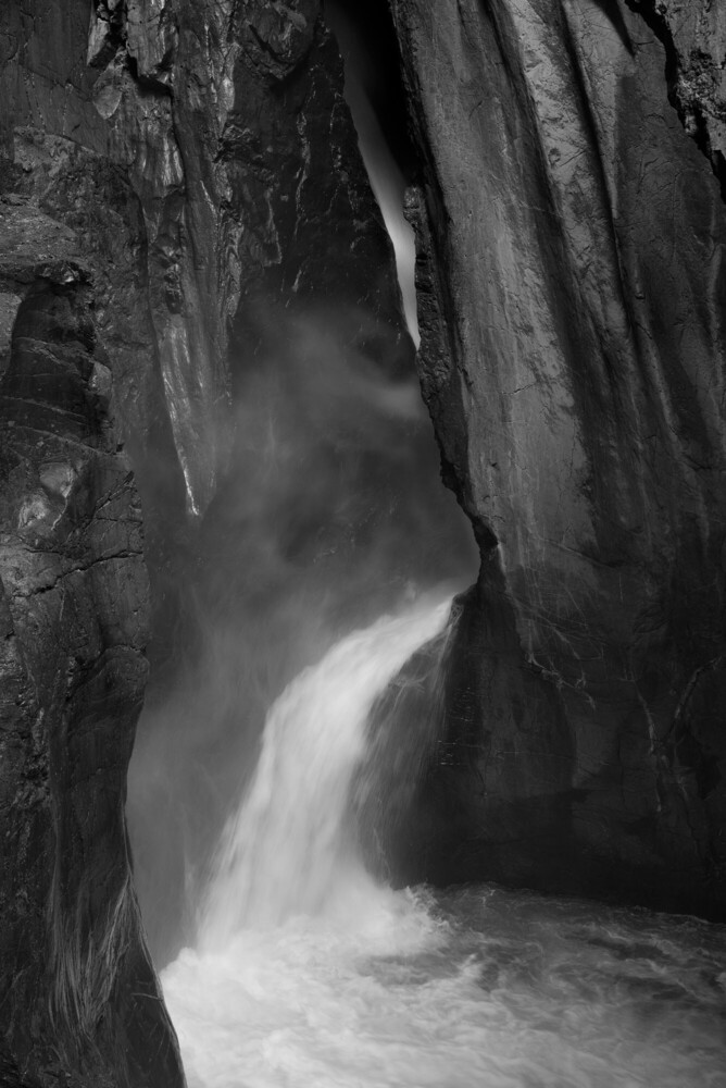 Black and white long exposure of a waterfall cascading through a narrow rock canyon, with mist rising against dark, textured stone walls.