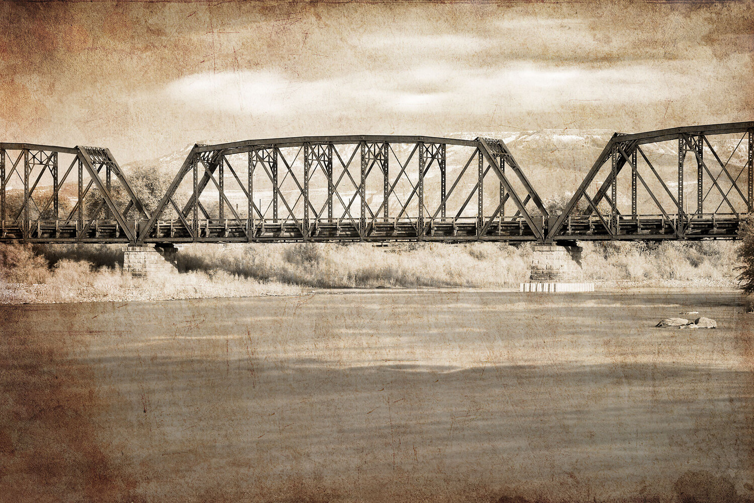 Sepia-toned steel truss bridge spanning a calm river beneath a cloudy sky, with dry grass along the banks.