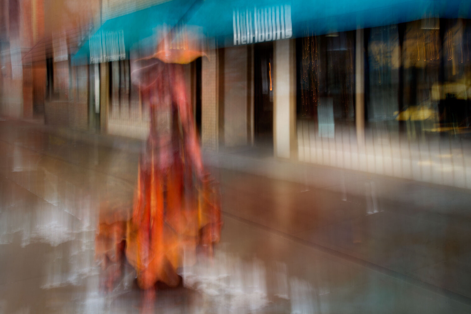 Blurred figure in a flowing orange-red dress standing on wet pavement beneath a teal awning on a city street