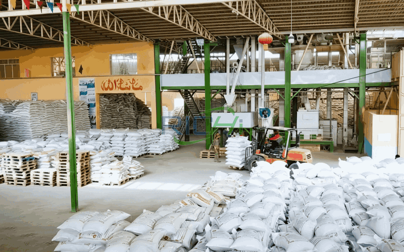 Warehouse interior with stacked white sacks on pallets, an orange forklift, and milling machinery on a mezzanine.