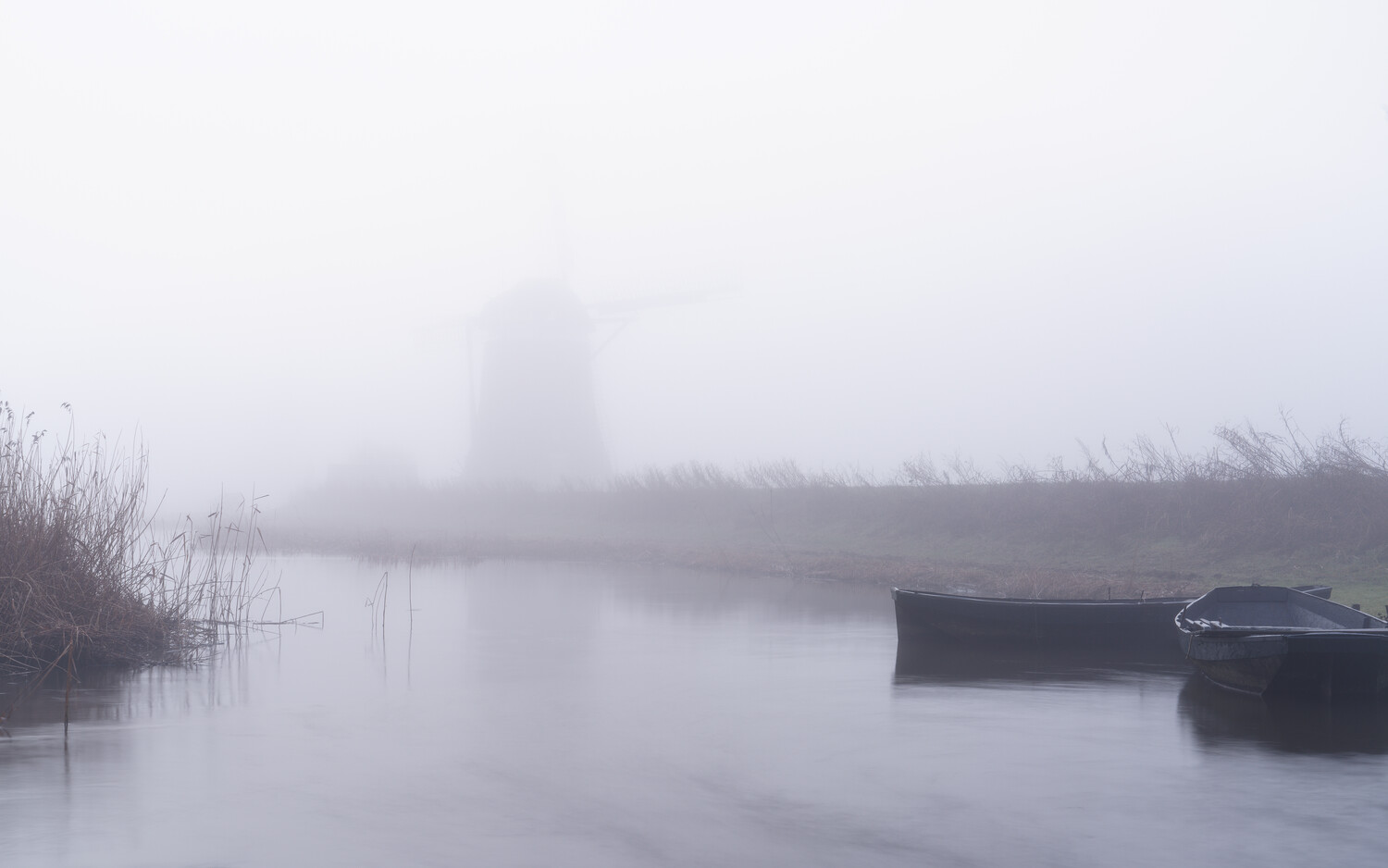 Dense fog veils a windmill on a still canal, with dark rowboats moored at right and reeds along the left bank.