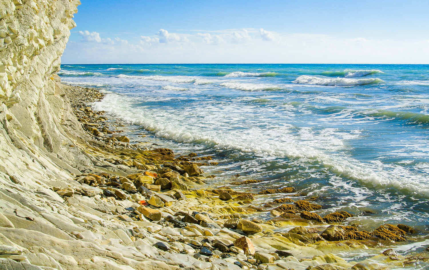 Chalk-white cliff beside a rocky shore, with waves breaking over algae-covered stones under a pale blue sky.