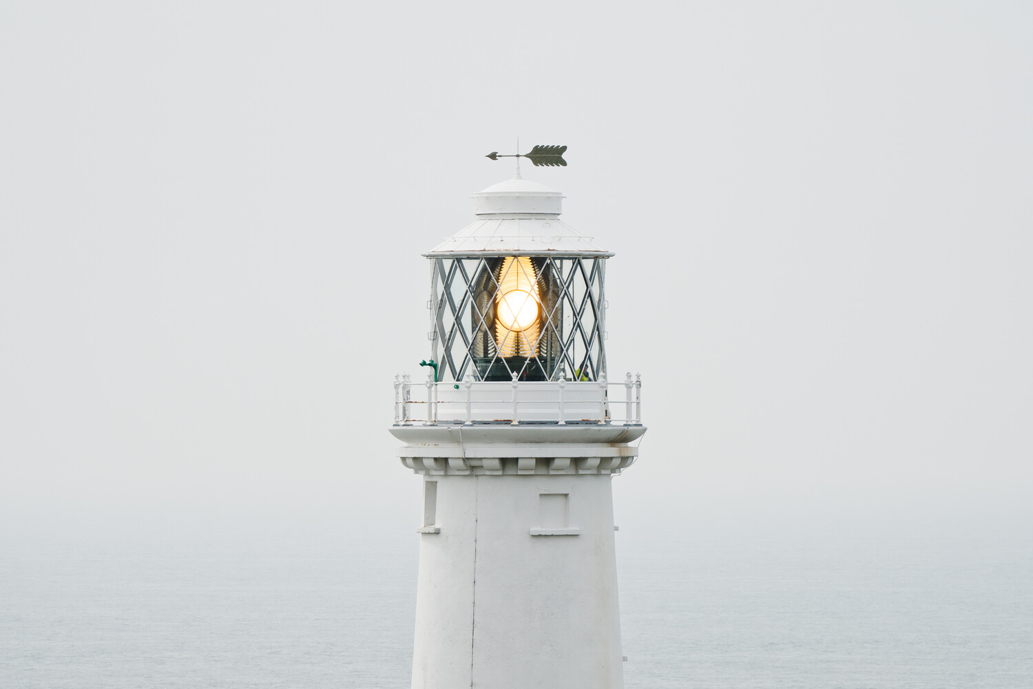 White lighthouse tower with a glowing lantern room against a pale, foggy sky and calm sea