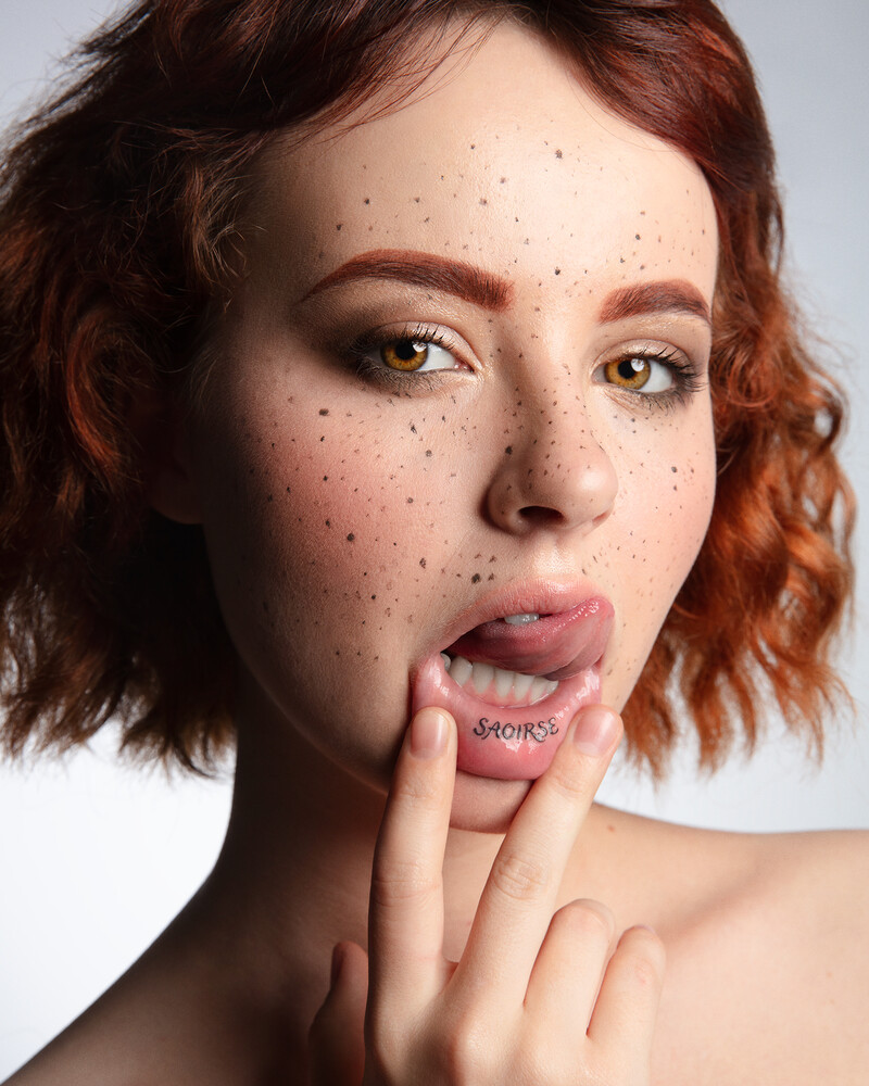 Portrait of a woman with reddish-brown wavy hair and freckles, touching her face with her finger