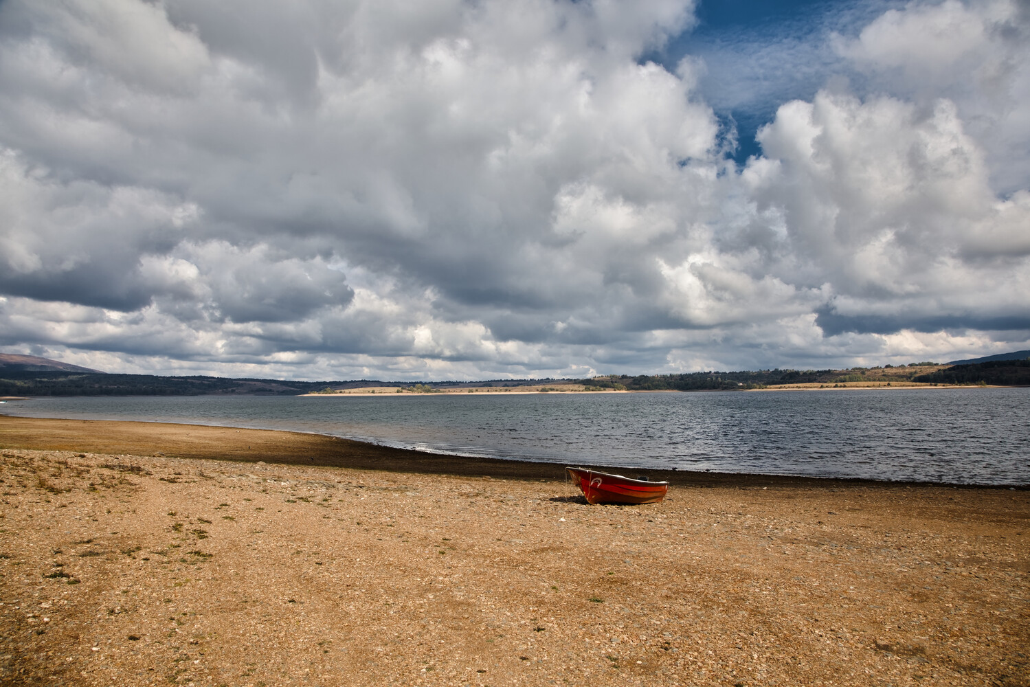 Beached red rowboat on sandy shore with calm water and dramatic cloud formations