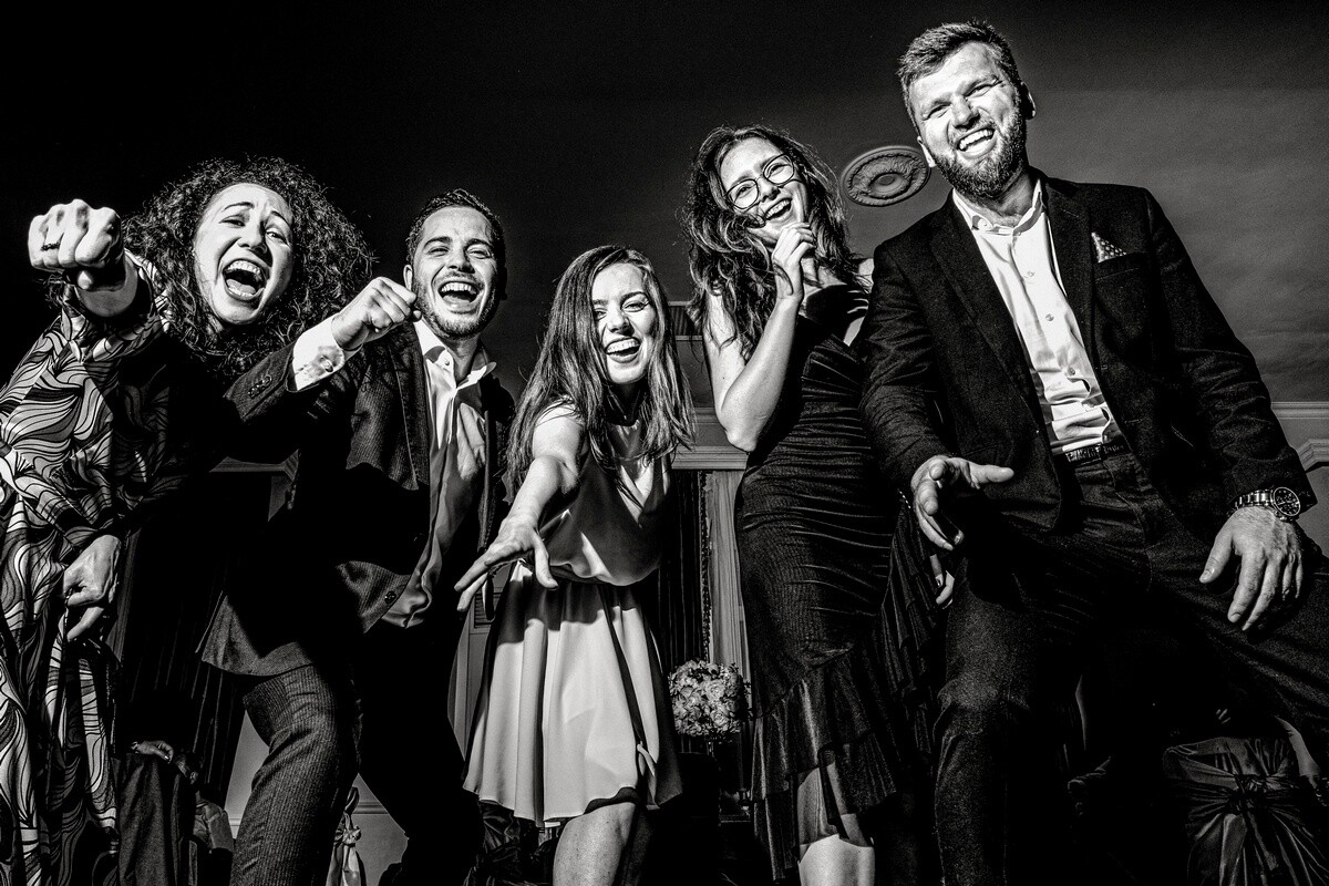 Six people in formal attire laughing and posing together against a dark background