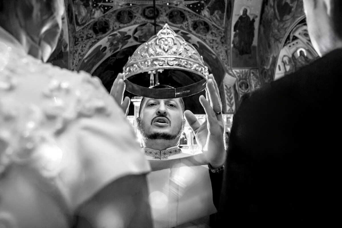 Man wearing ornate traditional headpiece in black and white, photographed among blurred figures and architectural details
