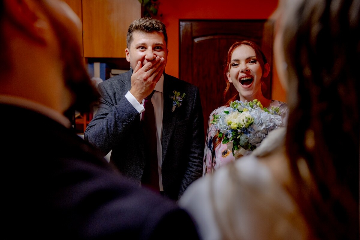 Groom laughing with hand to mouth while bride smiles beside him at wedding reception
