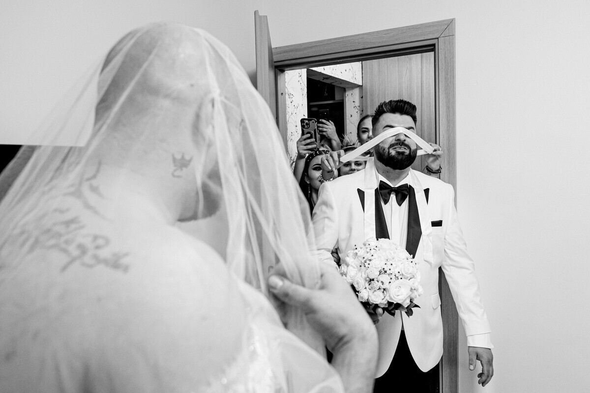 Groom in white jacket and black tie holding bouquet, seen through bride's veil in black and white