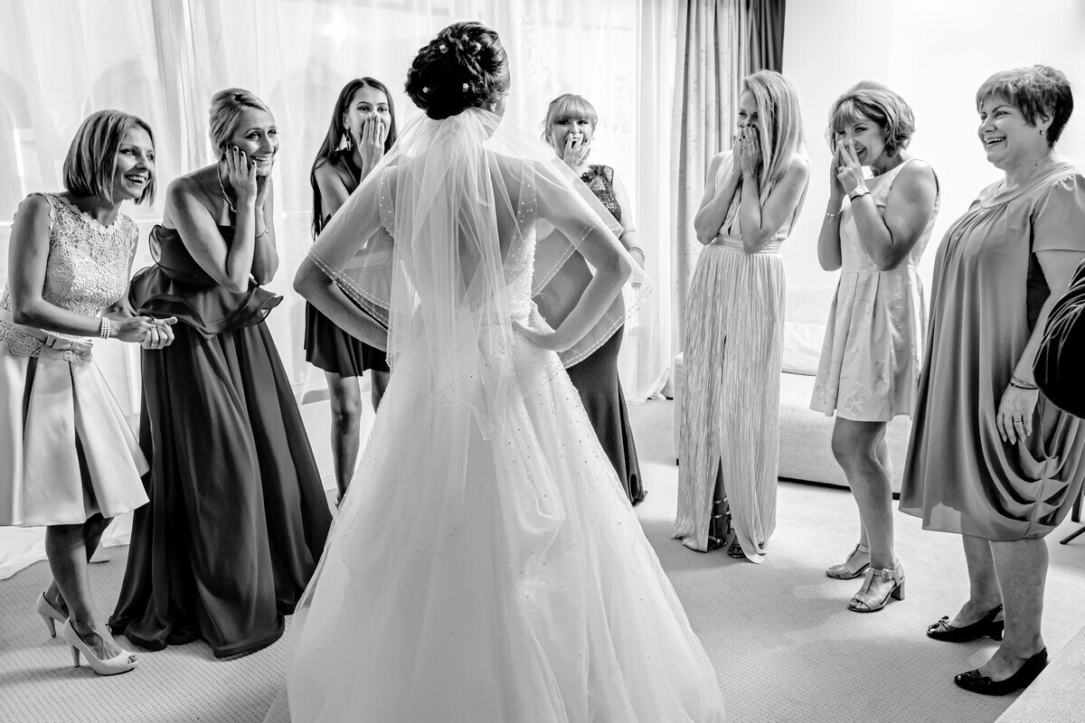 Bride in white wedding dress and veil surrounded by bridesmaids in various colored dresses during getting-ready moment