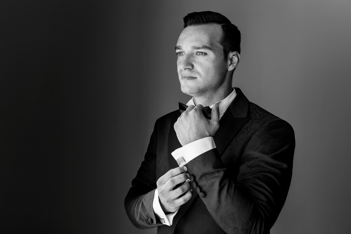 Man in formal black suit and bow tie adjusting his cufflinks while gazing thoughtfully to the side