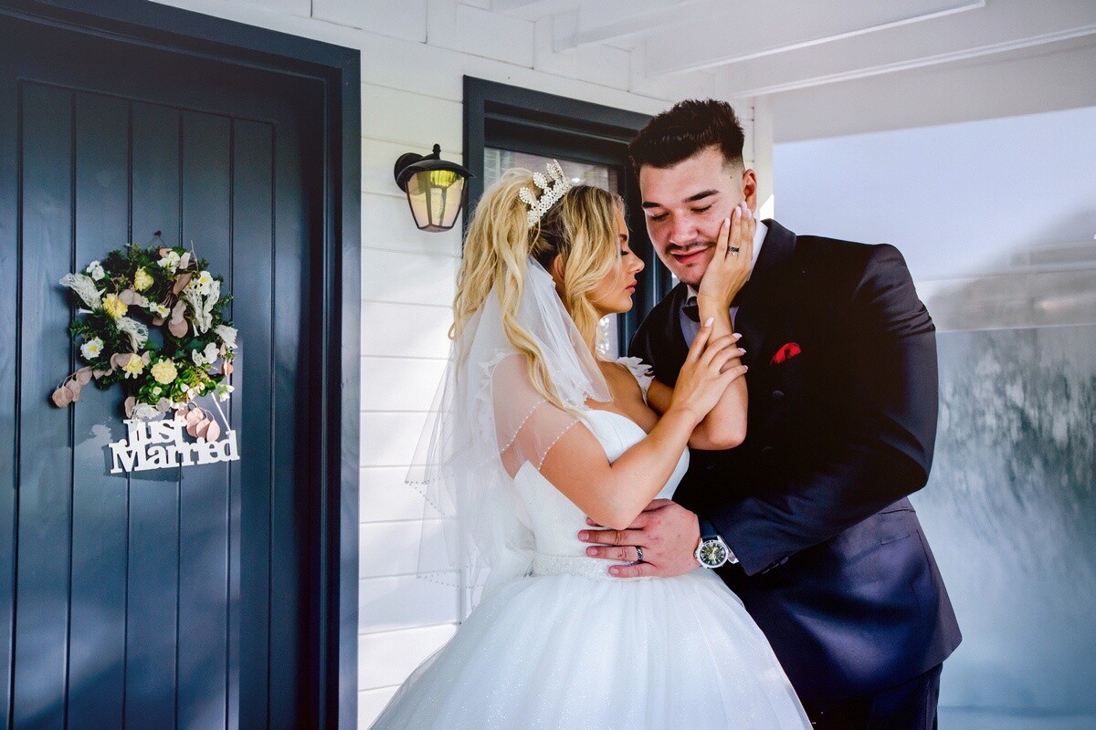Bride in a white gown and veil holding the face of a groom in a navy suit at a dark blue front door.