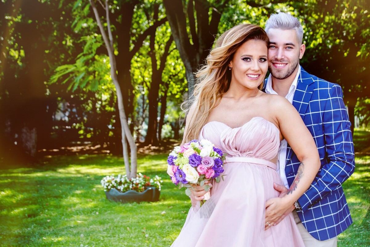Smiling couple embracing outdoors on a sunny lawn; the woman in a pale pink strapless gown holds a bouquet of purple and pink flowers.