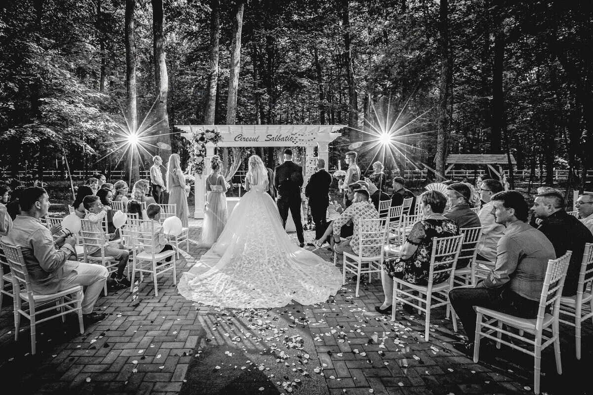 Black and white photo of a bride in a long lace gown with a cathedral train, facing a forest wedding ceremony under a white floral arch.