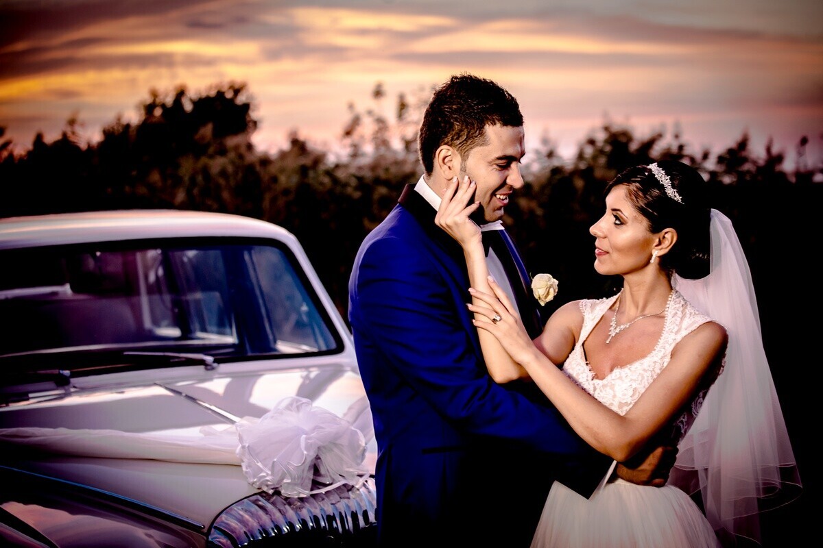 Bride and groom embracing at sunset beside a vintage white car, the groom in a navy suit and the bride in a lace gown with a veil.