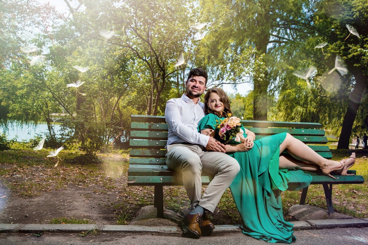 Bearded man in a white shirt and beige trousers sits on a green park bench with a smiling woman in a long teal dress holding a bouquet, birds flying around them.