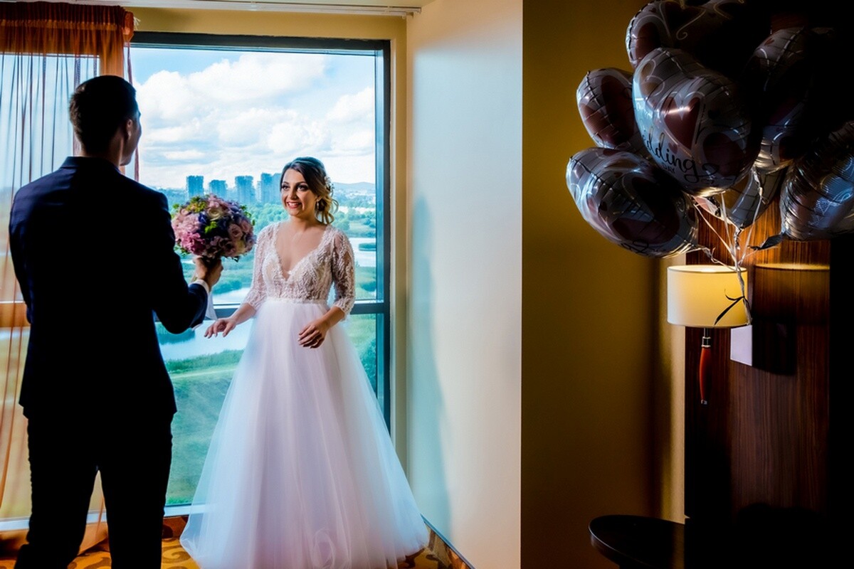 Smiling bride in a lace-sleeved white gown stands by a city-view window as a groom in a dark suit offers her a bouquet.