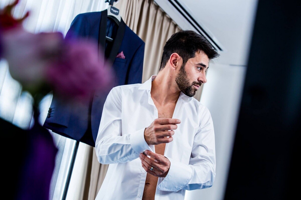 Man in an open white dress shirt buttoning his cuff, with a navy suit and pink pocket square hanging behind him