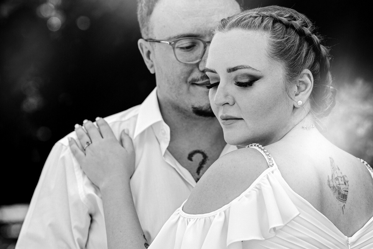 Black and white portrait of a bearded man in glasses embracing a woman with a braided updo, her shoulder bare, both gazing downward.
