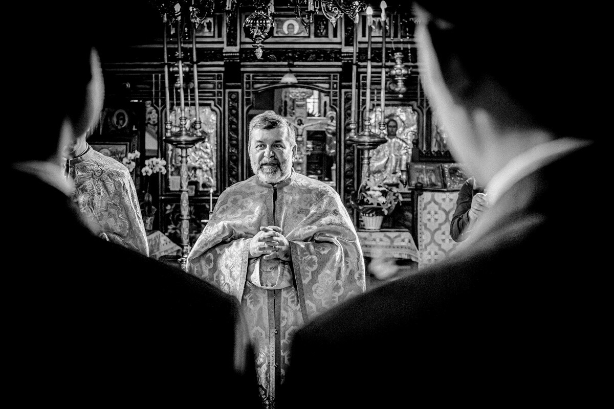 Smiling Orthodox priest in embroidered vestments with hands clasped, framed by blurred foreground figures in a church interior