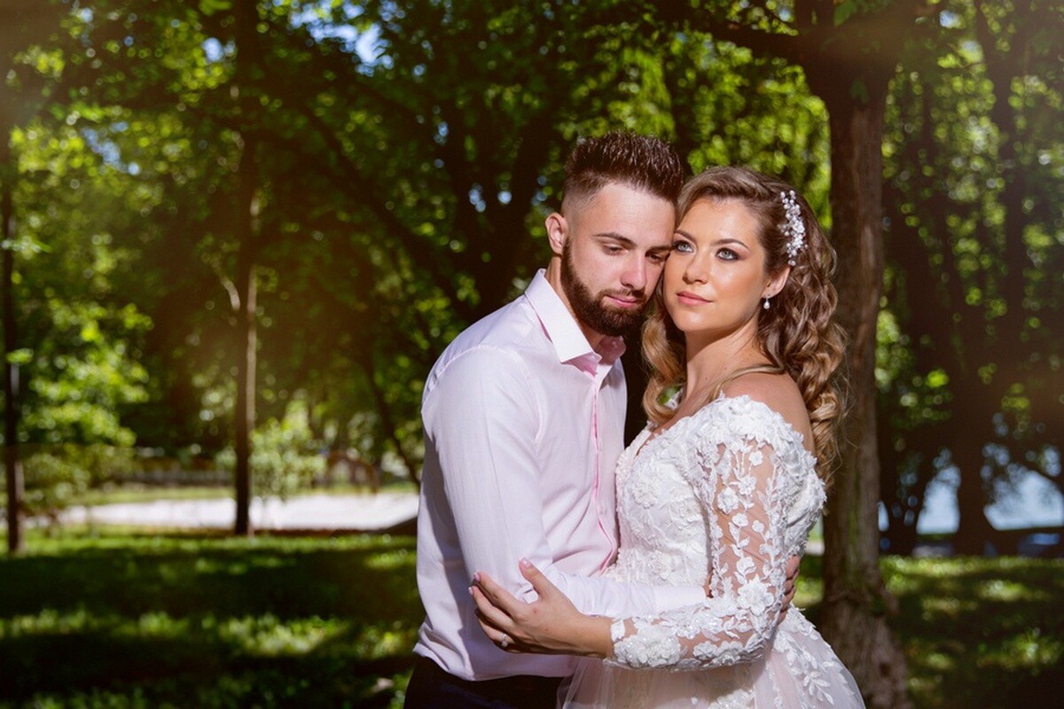 Bride in a white lace gown with long sleeves and a man in a pink dress shirt embracing outdoors among green trees on a sunny day.