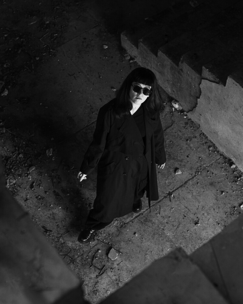 Person in dark coat and sunglasses standing in concrete stairwell, shot from above