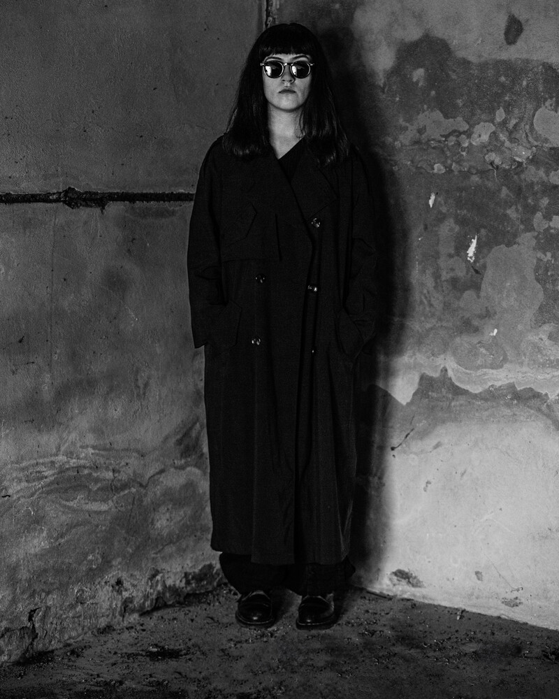 Person in long dark coat and sunglasses standing against weathered concrete wall