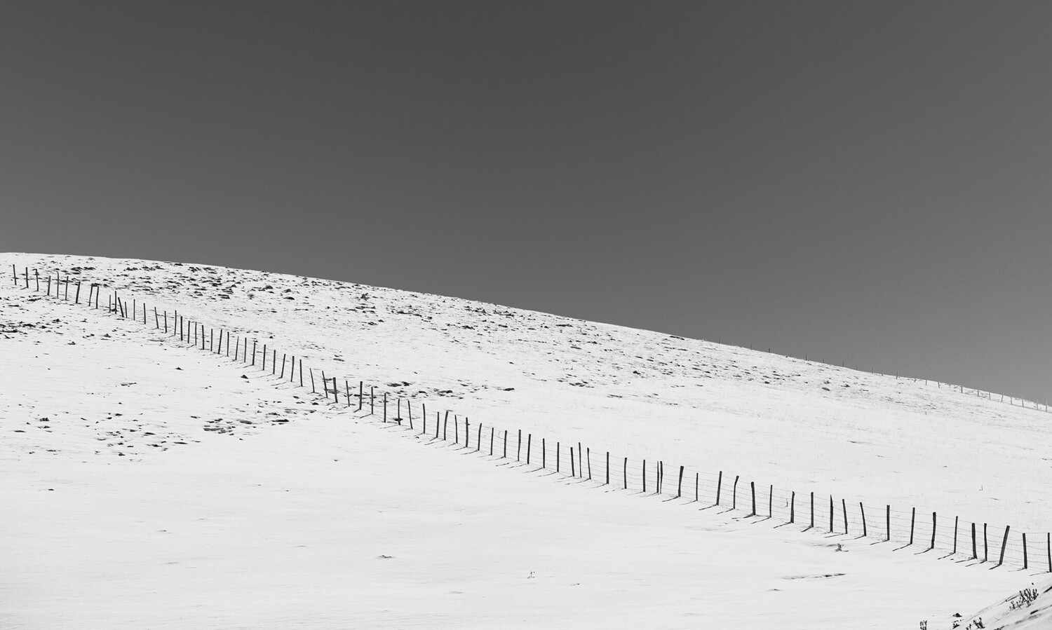 Snow-covered slope with wooden fence line running diagonally across the frame