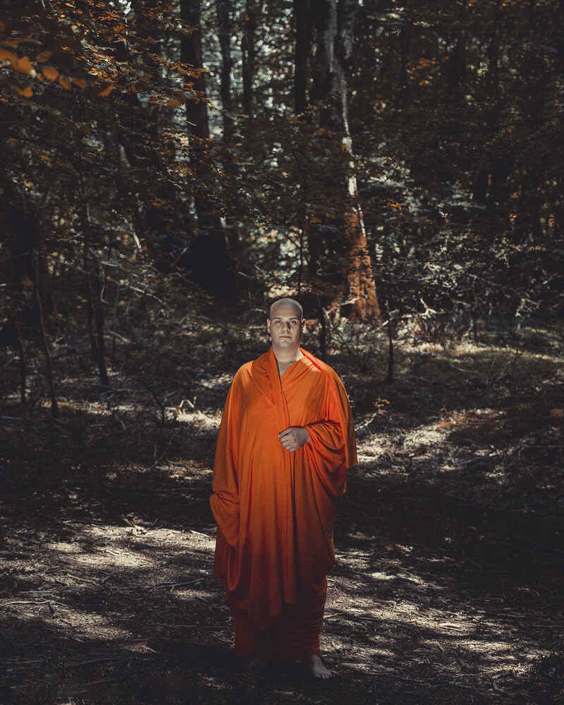 Monk in orange robes standing on a forest path with dappled sunlight