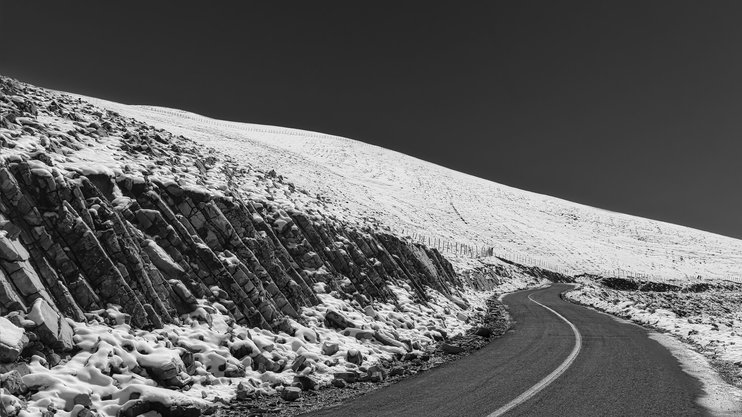 Winding asphalt road curves through snow-covered mountainous terrain under dark sky