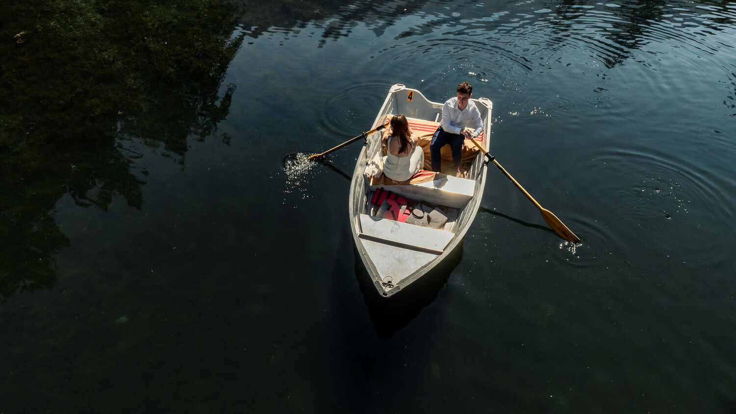 Two people rowing a white boat on calm, dark water with forested shoreline