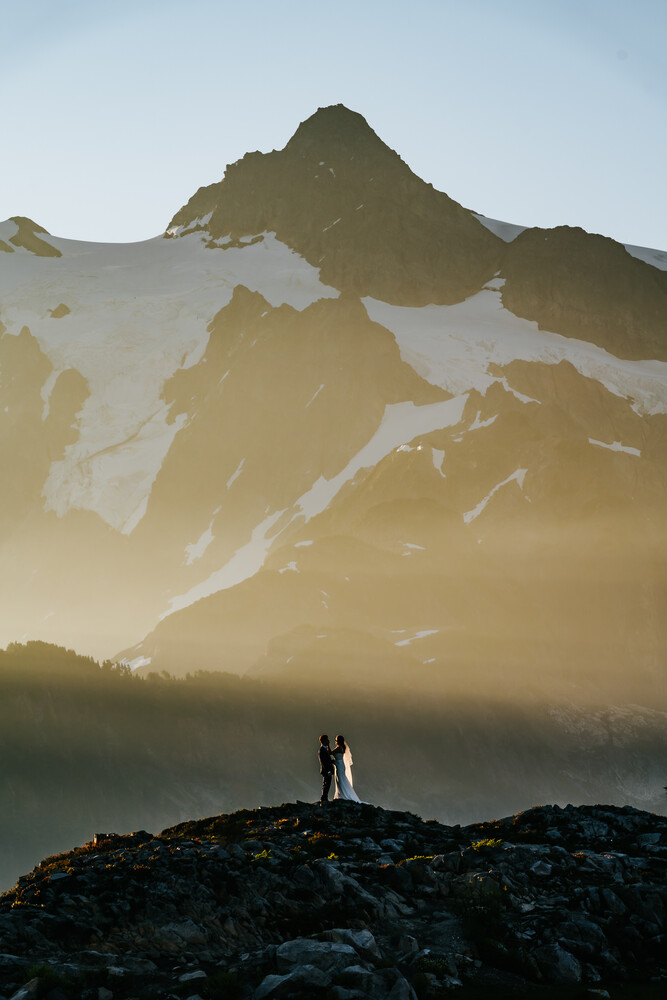 Solitary figure standing on rocky terrain beneath a snow-covered mountain peak shrouded in mist