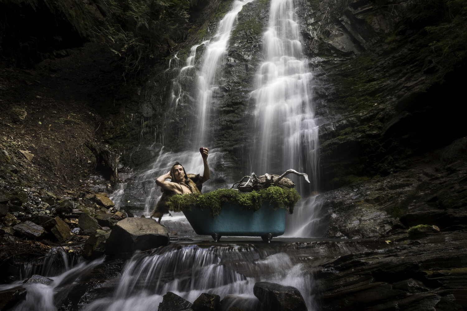 Woman in hiking gear posing on moss-covered rock beneath multi-tiered waterfall