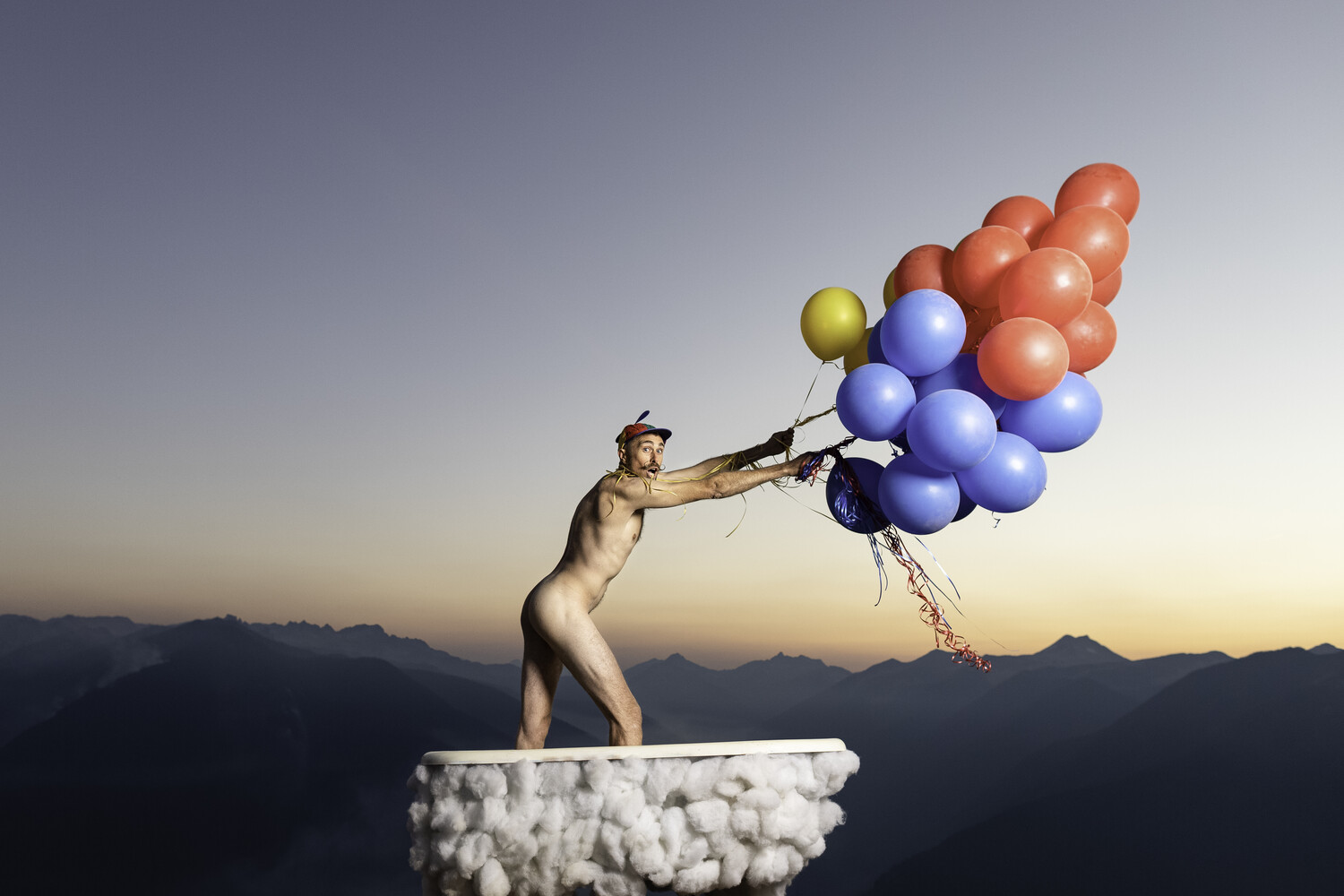 Nude figure standing on clouds holding bundle of colorful balloons against mountain landscape at sunset