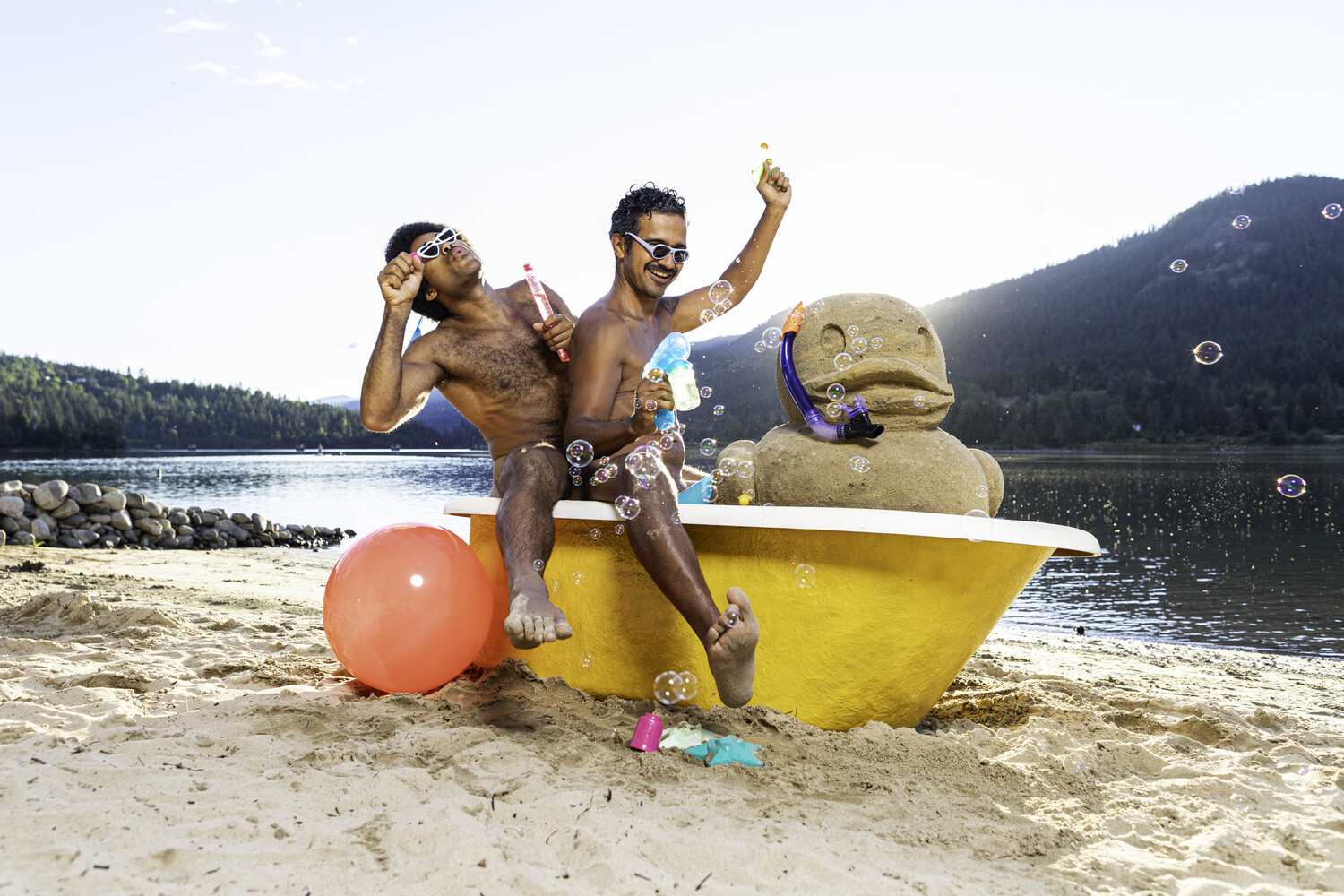 Two shirtless men posing playfully in a yellow bathtub on a beach, one wearing sunglasses with a fist raised