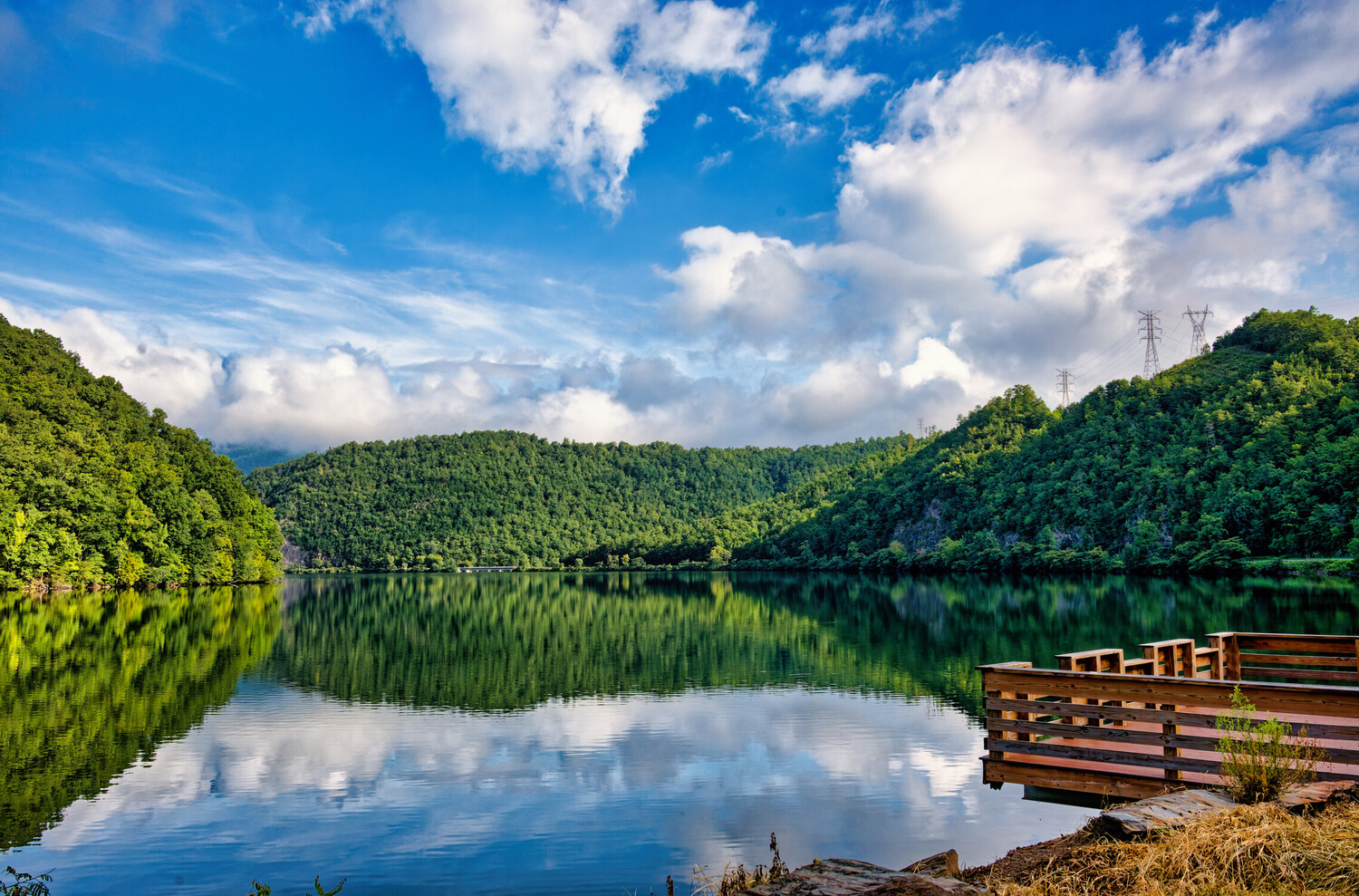 Serene lake surrounded by forested mountains with wooden dock in foreground and perfect mirror reflection