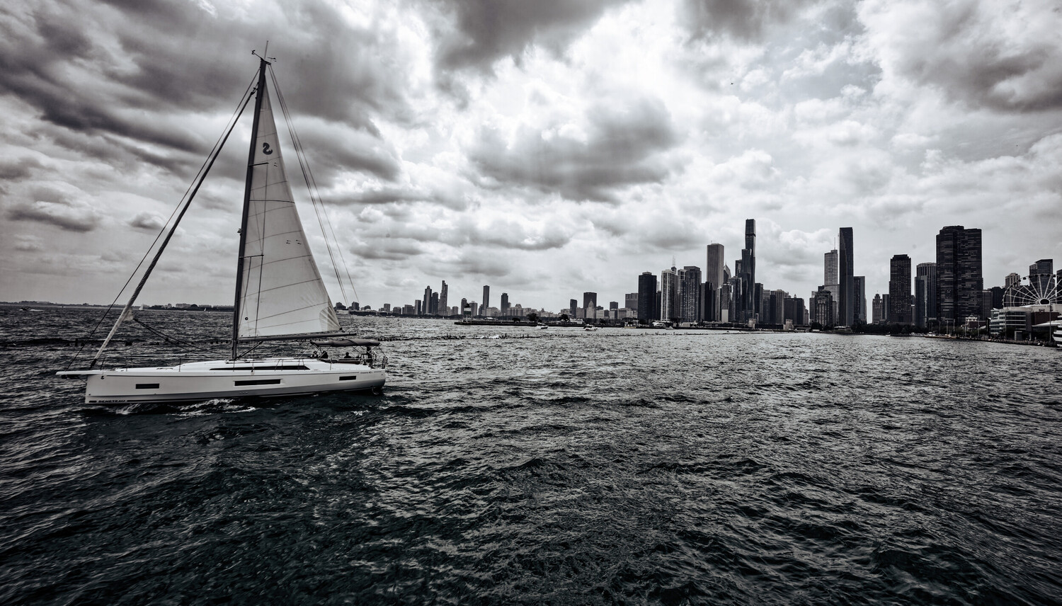 Sailboat with white sail on dark water facing a distant city skyline under dramatic cloudy sky