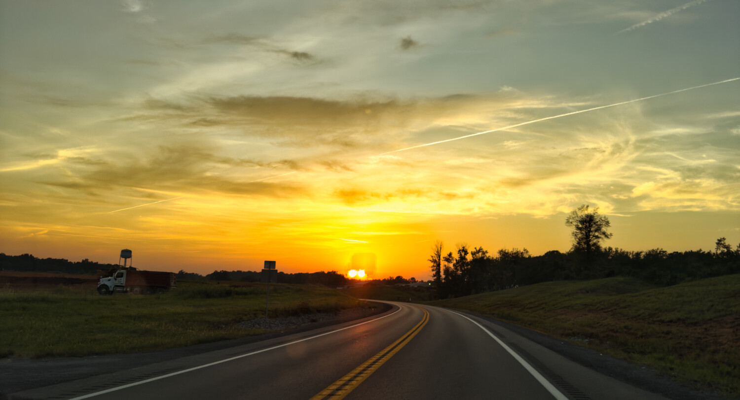 Highway stretching toward golden sunset with truck on left side and tree line on horizon