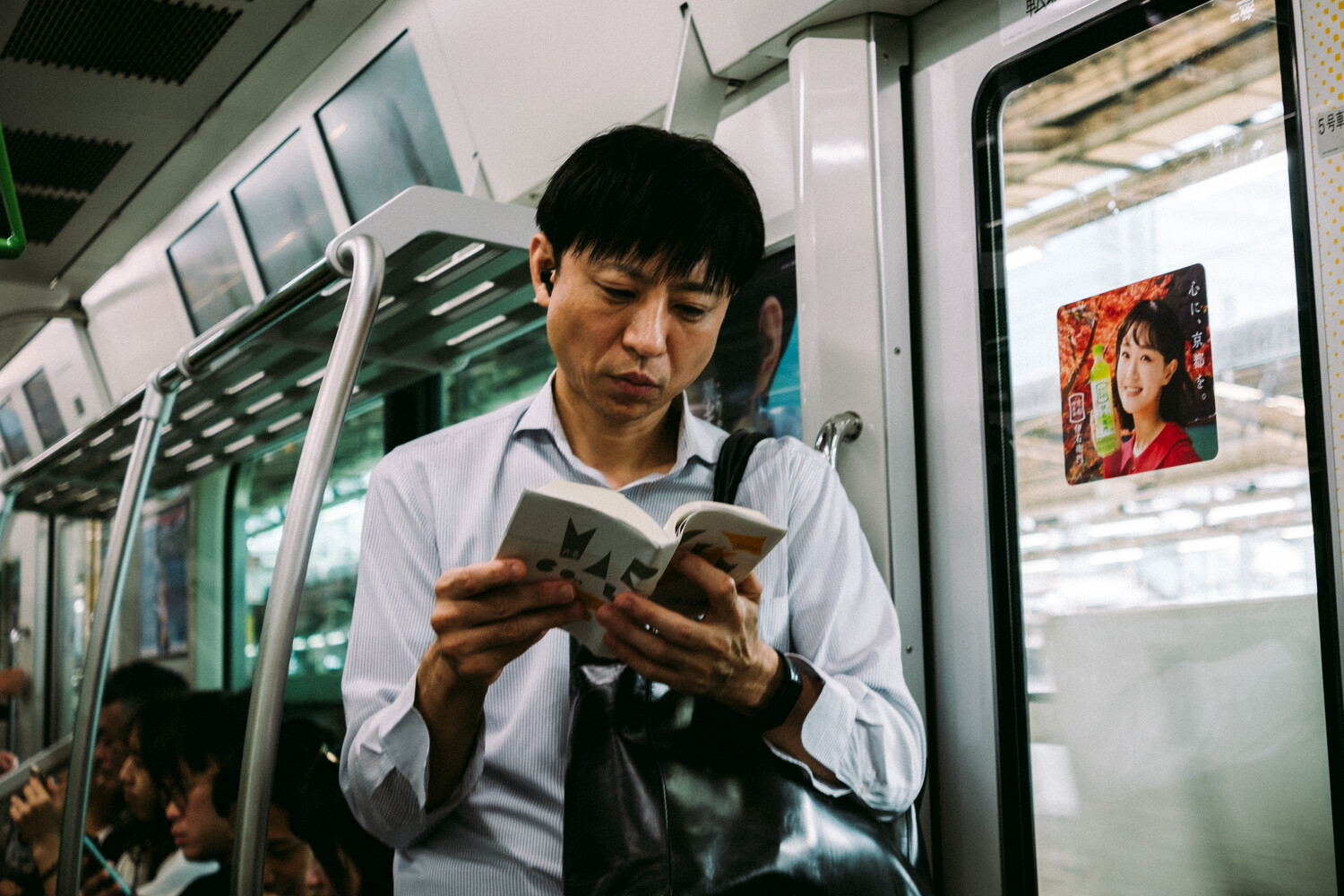 Man in white shirt reading a book on public transit, with colorful poster visible in window