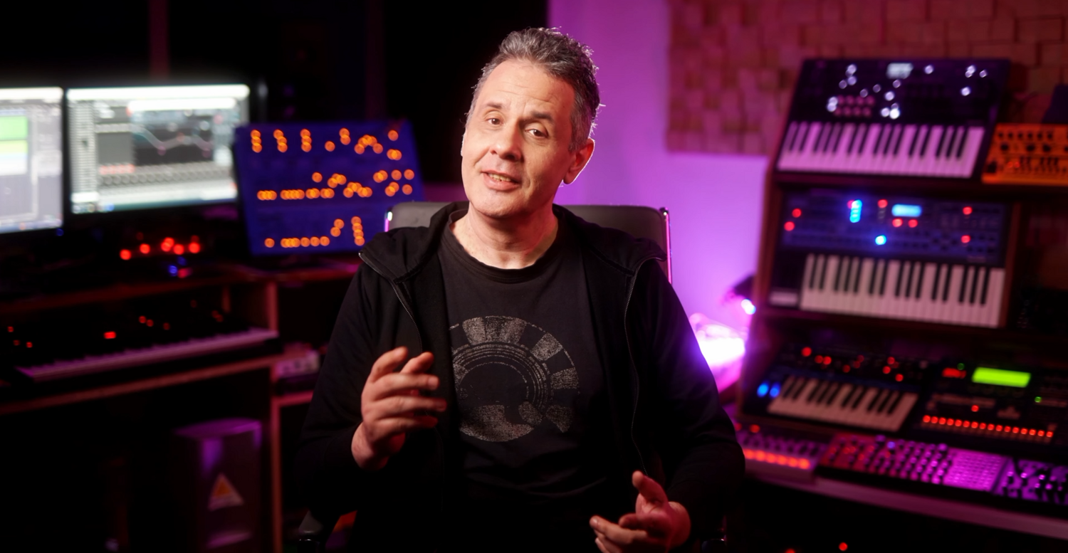 Man in black shirt gesturing while speaking in a purple-lit studio with synthesizers and music production equipment