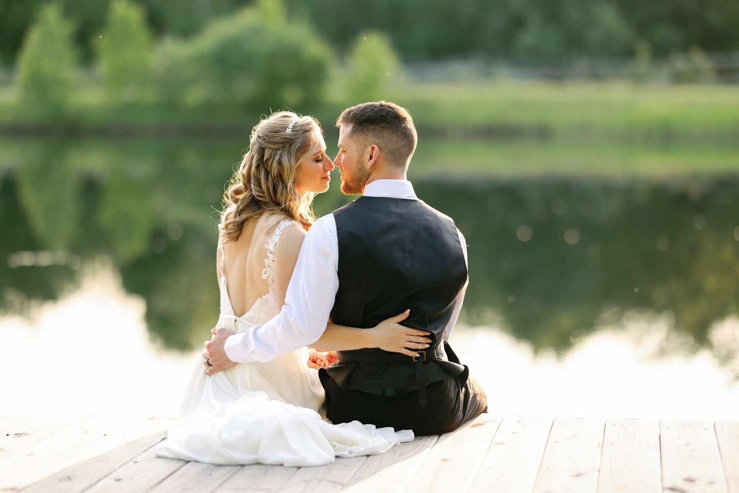 Couple in wedding attire sitting on a wooden dock facing each other with a calm river and green foliage behind them