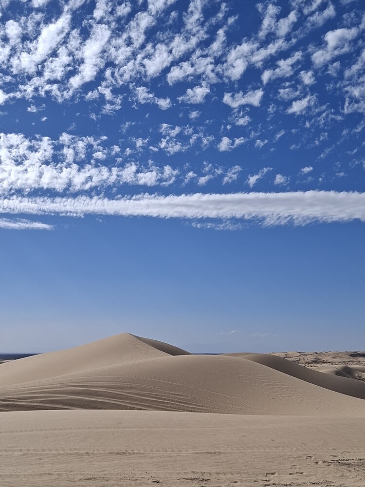 Sand dune under blue sky with wispy clouds and distant structures