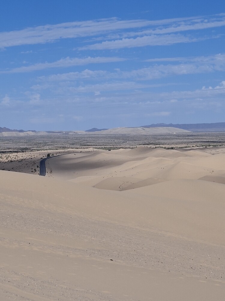 Person standing on expansive sand dunes with mountains visible on distant horizon under clear blue sky